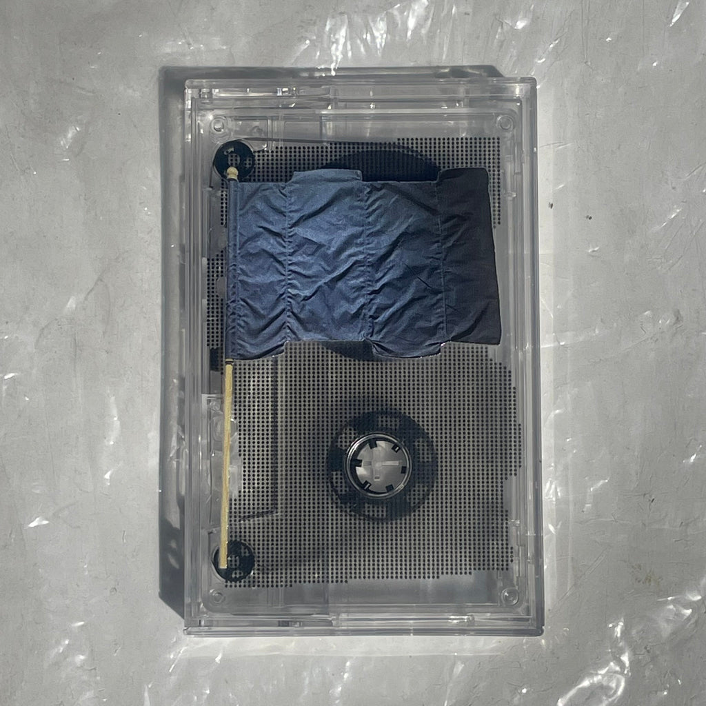A clear acrylic cassette-style display case containing a small blue puffer jacket and a wooden stick, photographed from above on a light gray wrinkled surface.