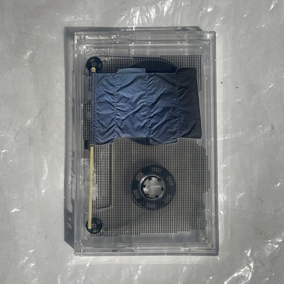 A clear acrylic cassette-style display case containing a small blue puffer jacket and a wooden stick, photographed from above on a light gray wrinkled surface.
